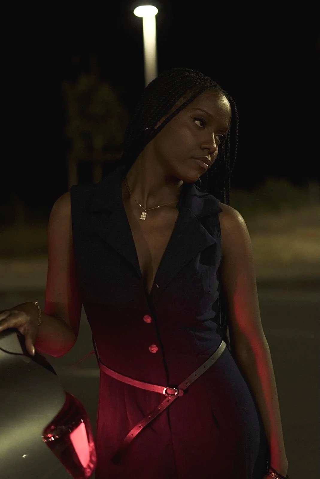 Une femme avec des cheveux tressés, portant un chemisier noir, une ceinture beige, et un collier doré, de nuit devant une voiture. Il y a un lampadaire en arrière-plan.