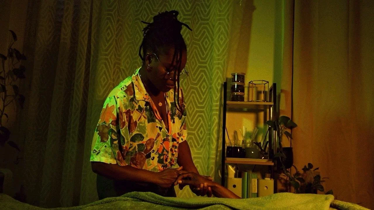 Une femme avec des cheveux en dreadlocks porte une chemise colorée et semble faire un massage ou une manipulation sur une autre personne dans une pièce avec un éclairage tamisé, des étagères avec des objets et des plantes visibles en arrière-plan.