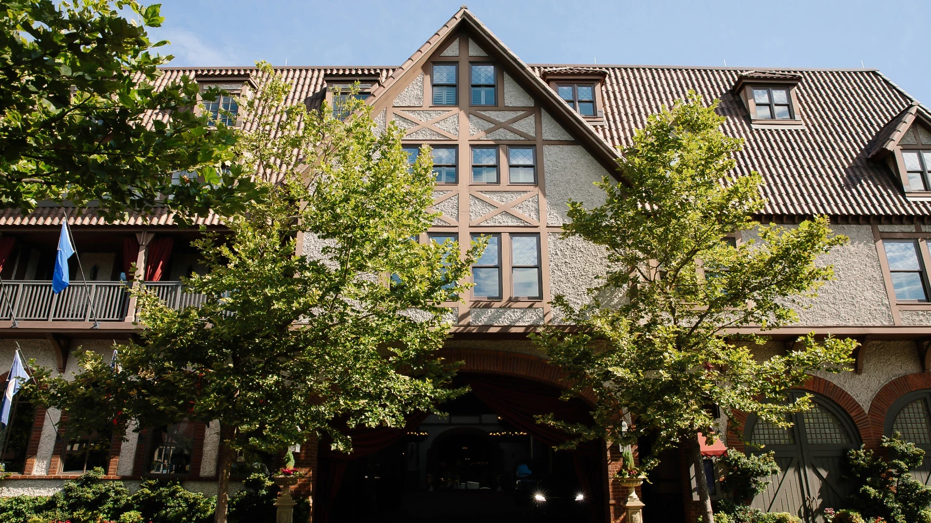 A multi-story building with a steep, tiled roof and decorative timber framing, surrounded by green trees and a clear blue sky.