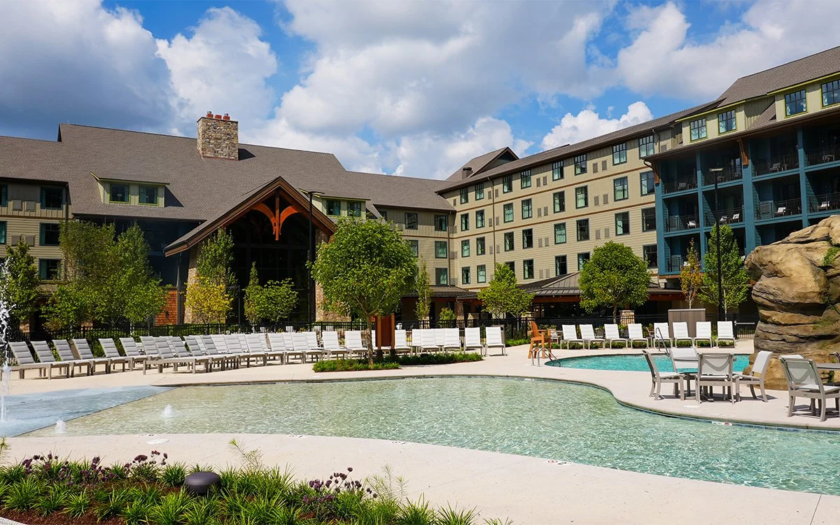 A hotel pool area with lounge chairs, tables, and a small pool surrounded by trees, with a large multi-story hotel building in the background under a partly cloudy sky.