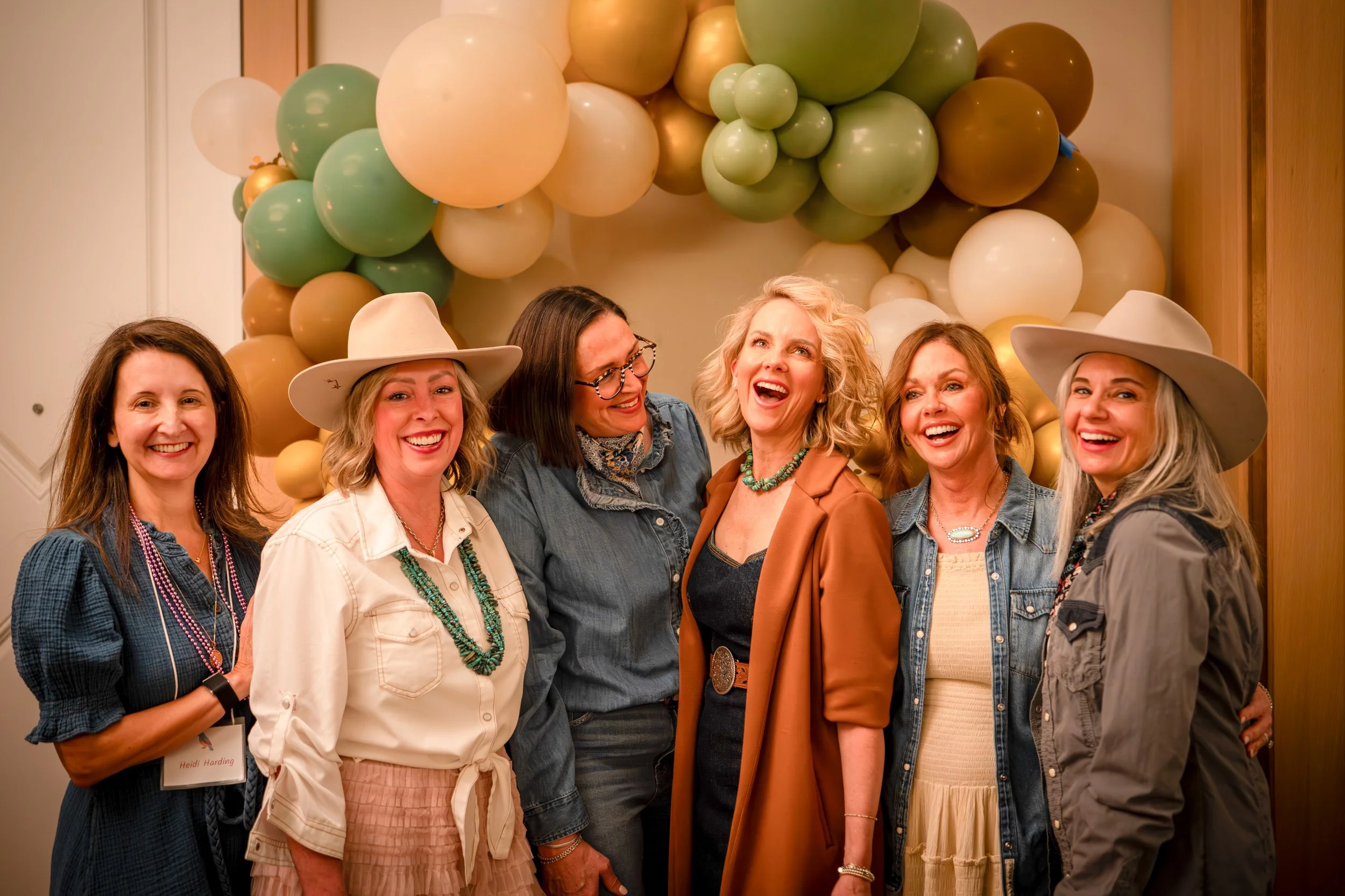 A group of six women smiling and standing close together in front of a backdrop of colorful balloons, some of which are gold, green, white, and beige. Two women are wearing large white hats, and the woman in the middle is laughing. They are indoors, 