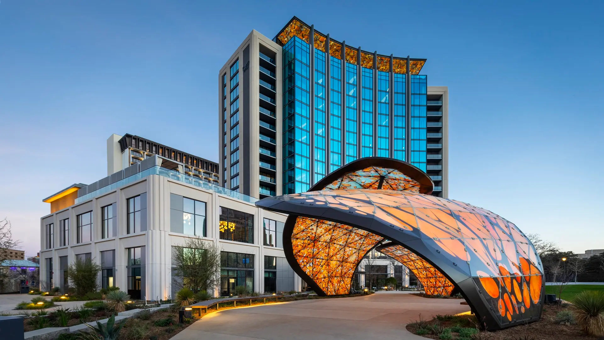 Modern multi-story building with blue glass windows and a metal and glass architectural structure in the foreground, featuring orange and yellow leaf patterns illuminated from within, set against a clear blue sky.