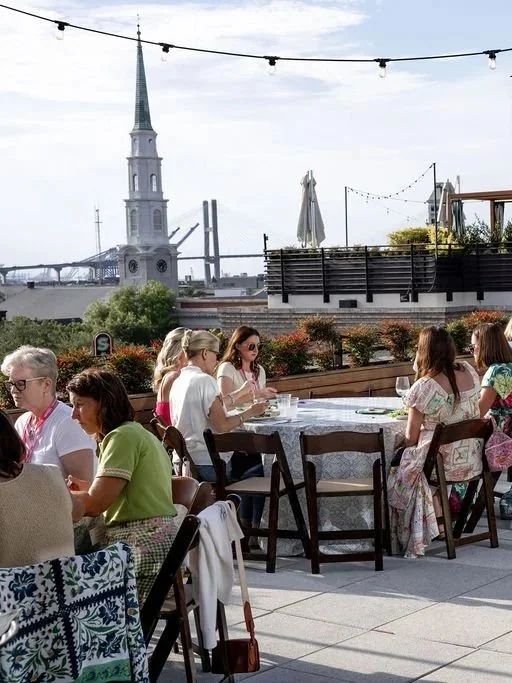 People gathered at an outdoor rooftop restaurant during the daytime, with several tables of mostly women dining and socializing, overlooking a cityscape with a church steeple and bridge in the background, and string lights overhead.