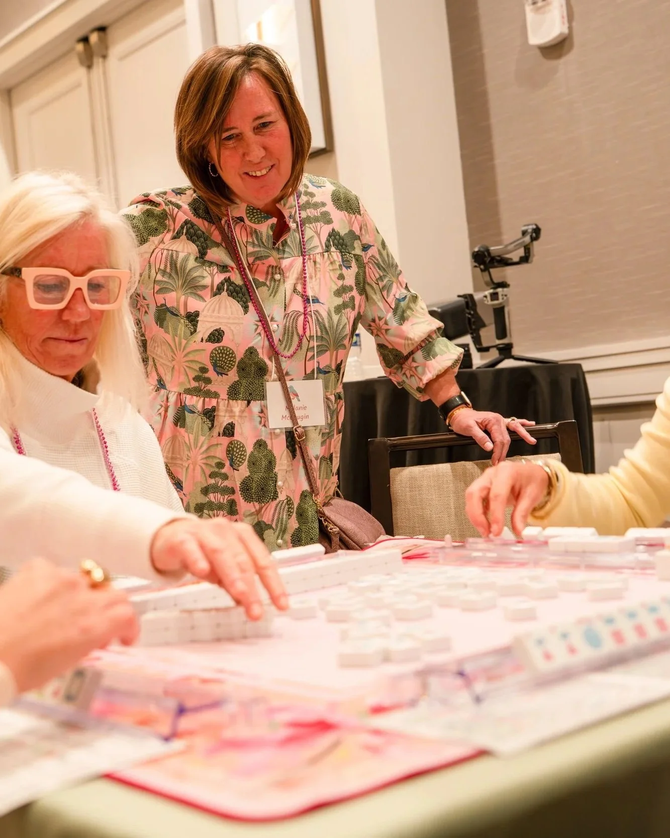 Four women are seated at a table playing a game of American Mahjo, with one woman standing and observing. The women are dressed in colorful clothing and have name tags. The setting appears to be a conference or social event in a large, well-lit room.