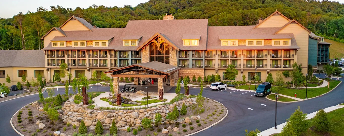 A large multi-story hotel surrounded by a landscaped parking lot and greenery, with forests on a hillside in the background.