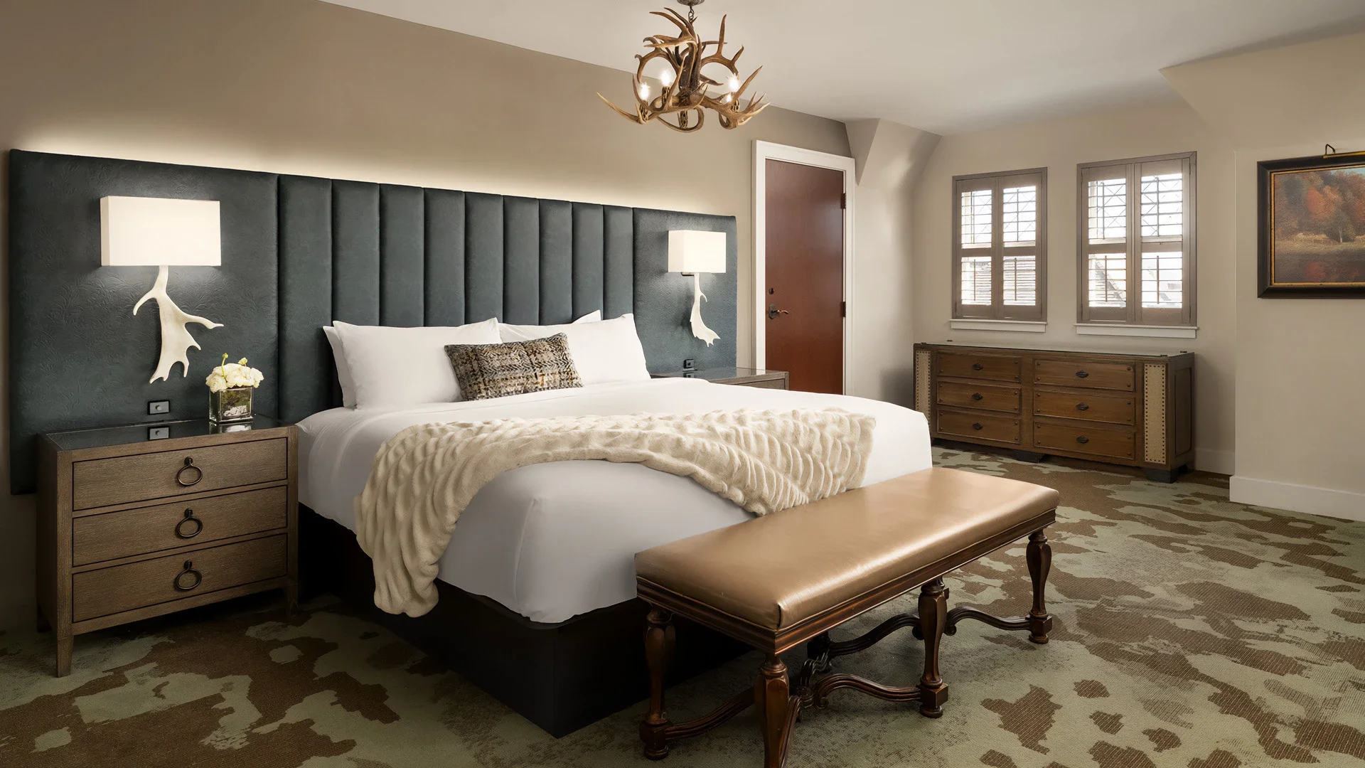 A bedroom with a large bed with white bedding and a furry blanket, dark headboard, and wooden furniture, including nightstands, dresser, and bench, with decorative antler-shaped wall lamps, a chandelier, and framed artwork, natural light from windows, and a patterned rug.
