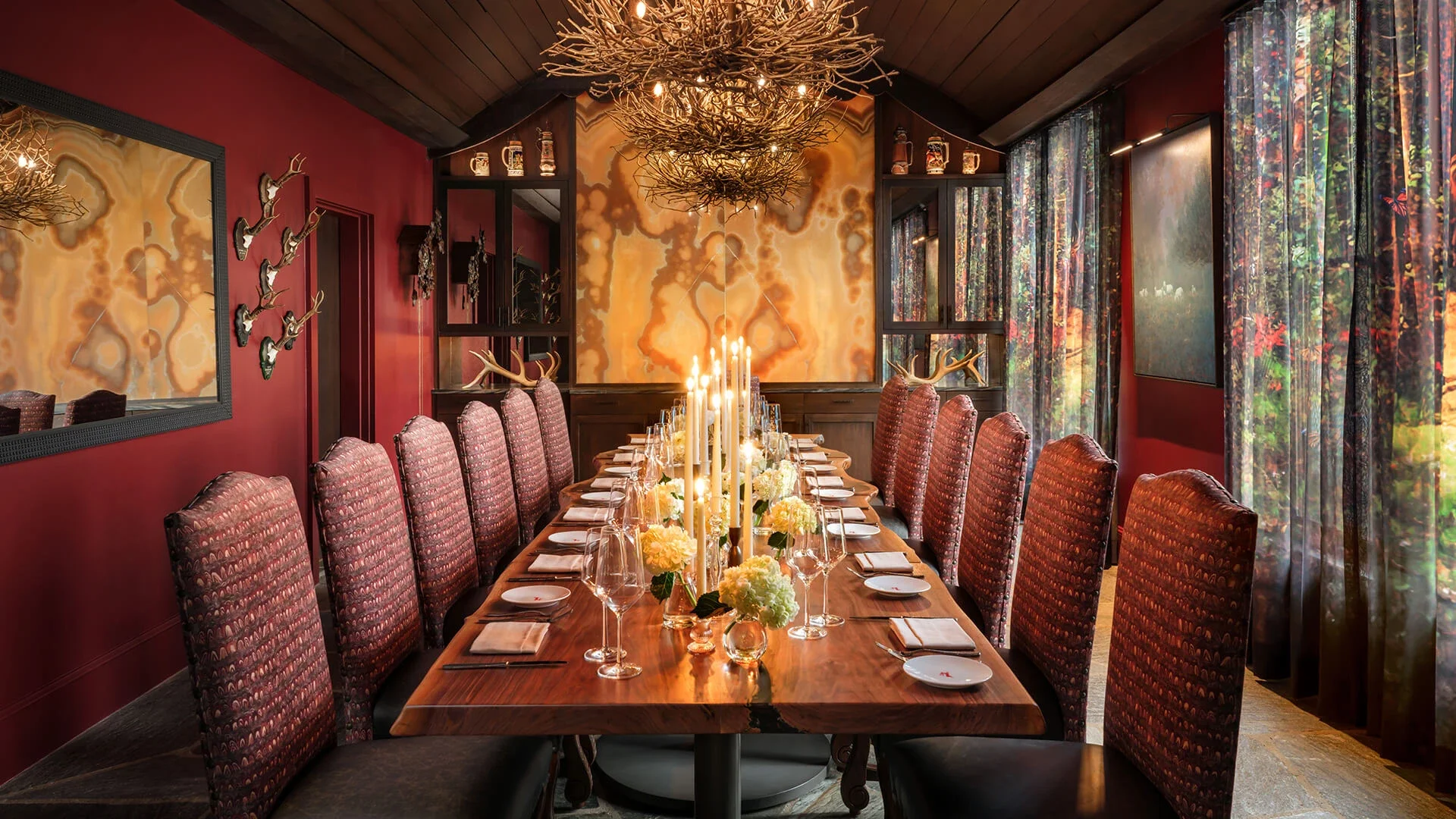 A dining room with a long wooden table set with white plates, wine glasses, and floral centerpieces. The chairs are upholstered in a patterned fabric, and the walls are painted deep red. Decorative antler fixtures are on the walls, and a chandelier made of intertwined branches hangs from the wooden vaulted ceiling. Large windows with colorful floral curtains let in natural light.