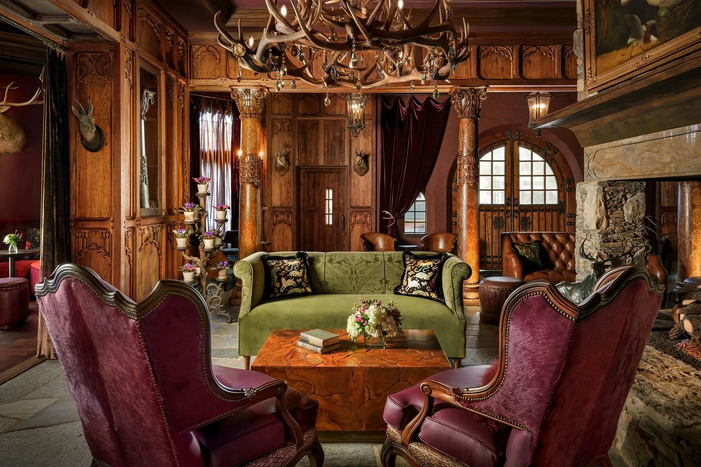 A luxurious living room with wood-paneled walls, a stone fireplace, velvet and leather chairs, vintage decor, and mounted deer heads, creating a rustic yet elegant atmosphere.