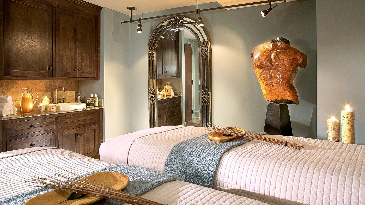 A bedroom with two beds, a large decorative mirror, a sculpture of a human torso on a stand, candles, and wooden cabinets.