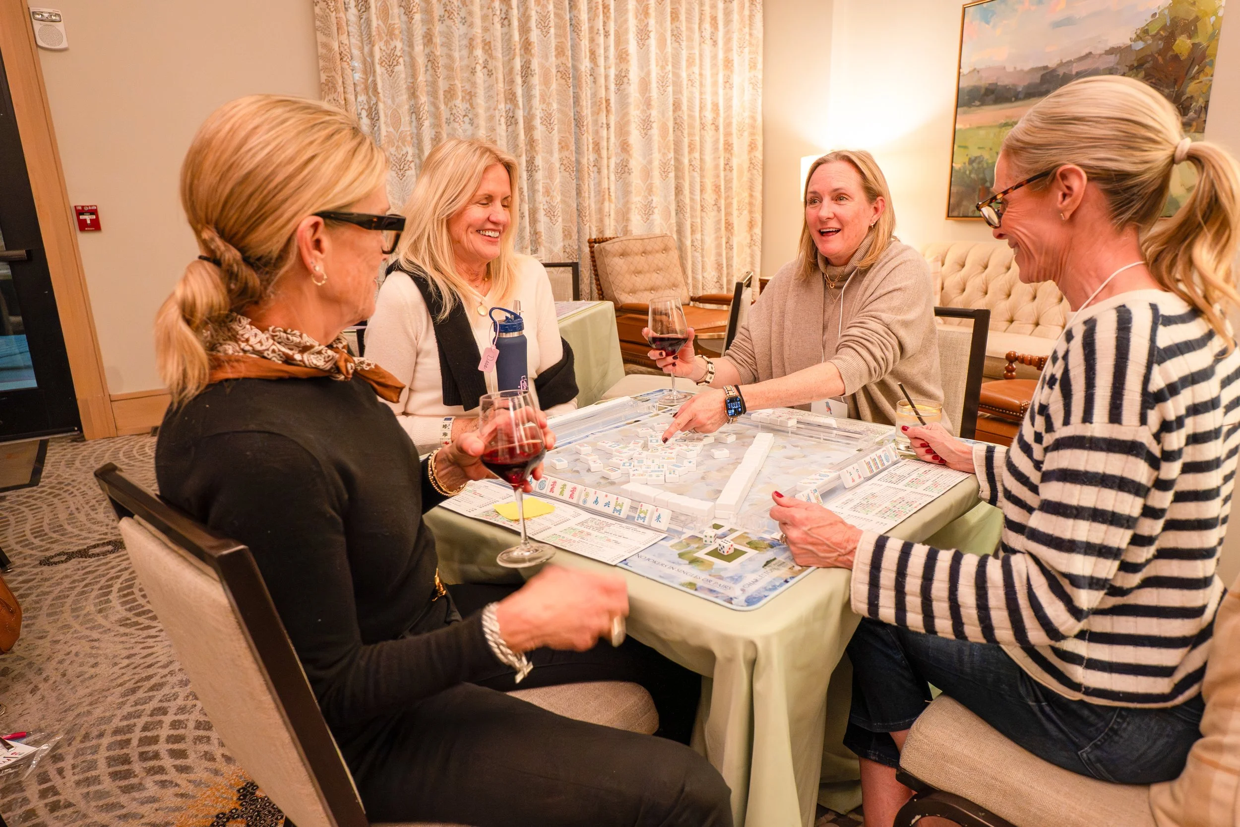 Four women sitting around a table playing a board game, with drinks in their hands, smiling and engaging in conversation in a warmly lit room.