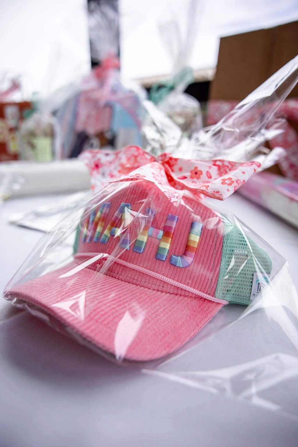 Pink Mahj baseball cap with colorful embroidered letters, wrapped in clear plastic and decorated with pink floral ribbon, on a white surface.