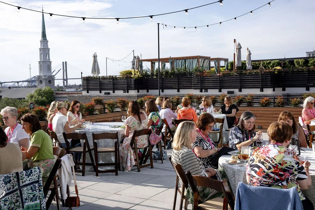 People gathered at an outdoor rooftop restaurant during the daytime, with several tables of mostly women dining and socializing, overlooking a cityscape with a church steeple and bridge in the background, and string lights overhead.