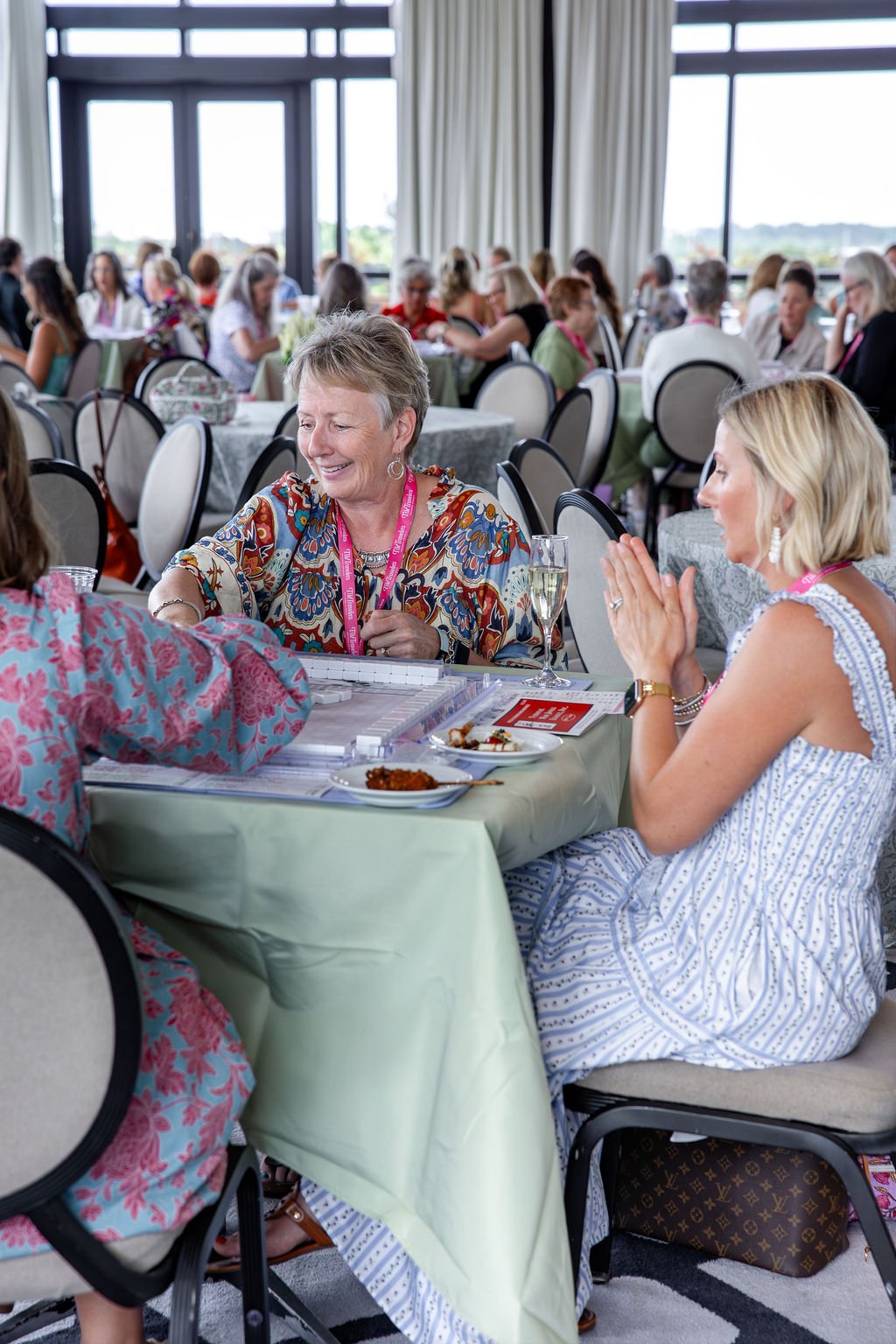 Women sitting at a banquet table during a social gathering, chatting and smiling with food and drinks in front of them, in a bright room with large windows.