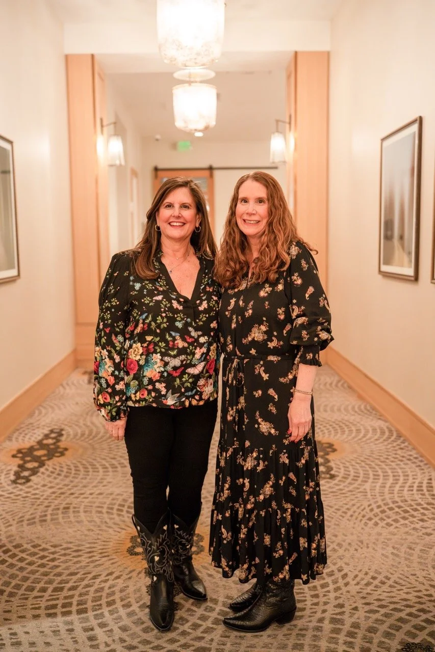 Two women standing in a hallway with framed pictures on the walls, smiling at the camera. They are dressed in floral patterned outfits, with one in black pants and cowboy boots, and the other in a black dress with boots.