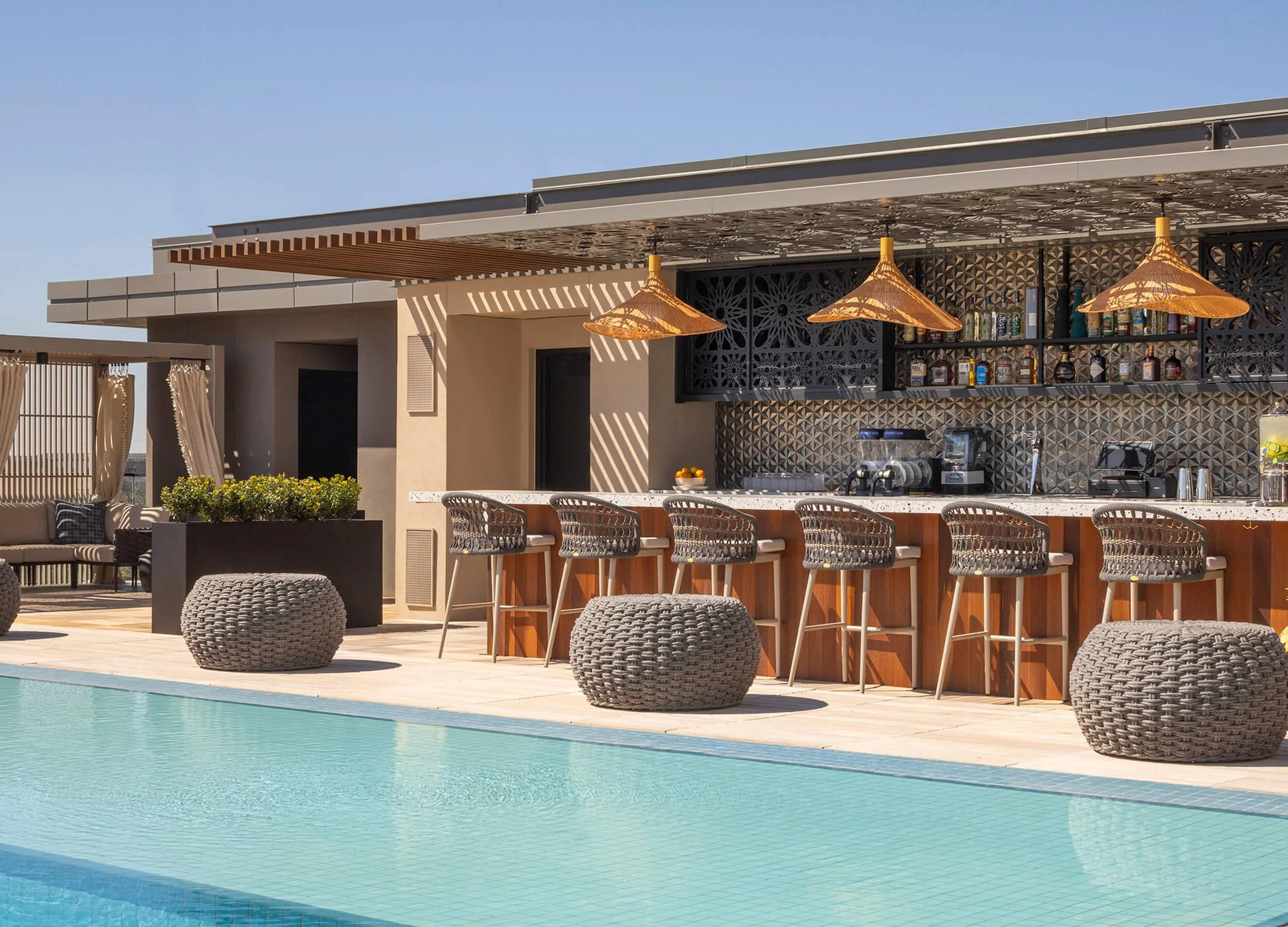 A modern outdoor poolside bar area with a countertop, barstools, and hanging bamboo pendant lights, overlooking a swimming pool, with a clear blue sky in the background.
