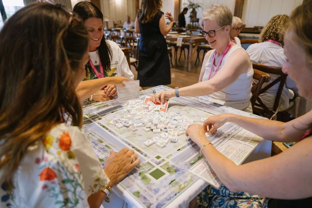 Four women are sitting at a table playing a game with American Mahjong tiles, smiling and interacting in a well-lit room with other people in the background.