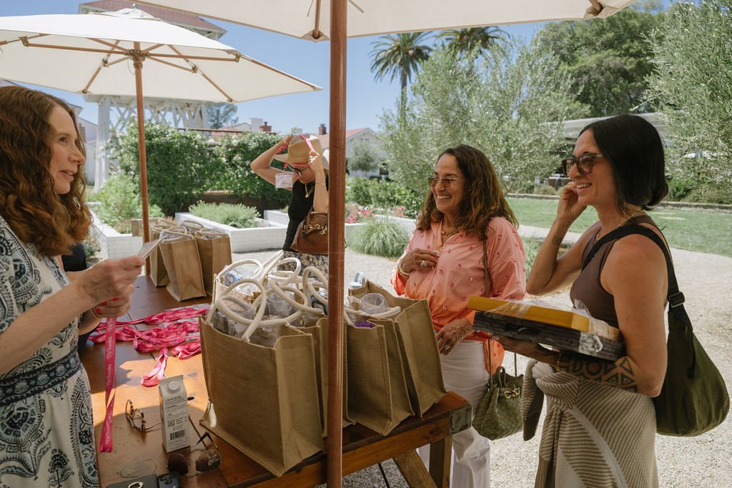 Three women talking and laughing at an outdoor market stall with tote bags and supplies, under umbrellas, with trees and a blue sky in the background.