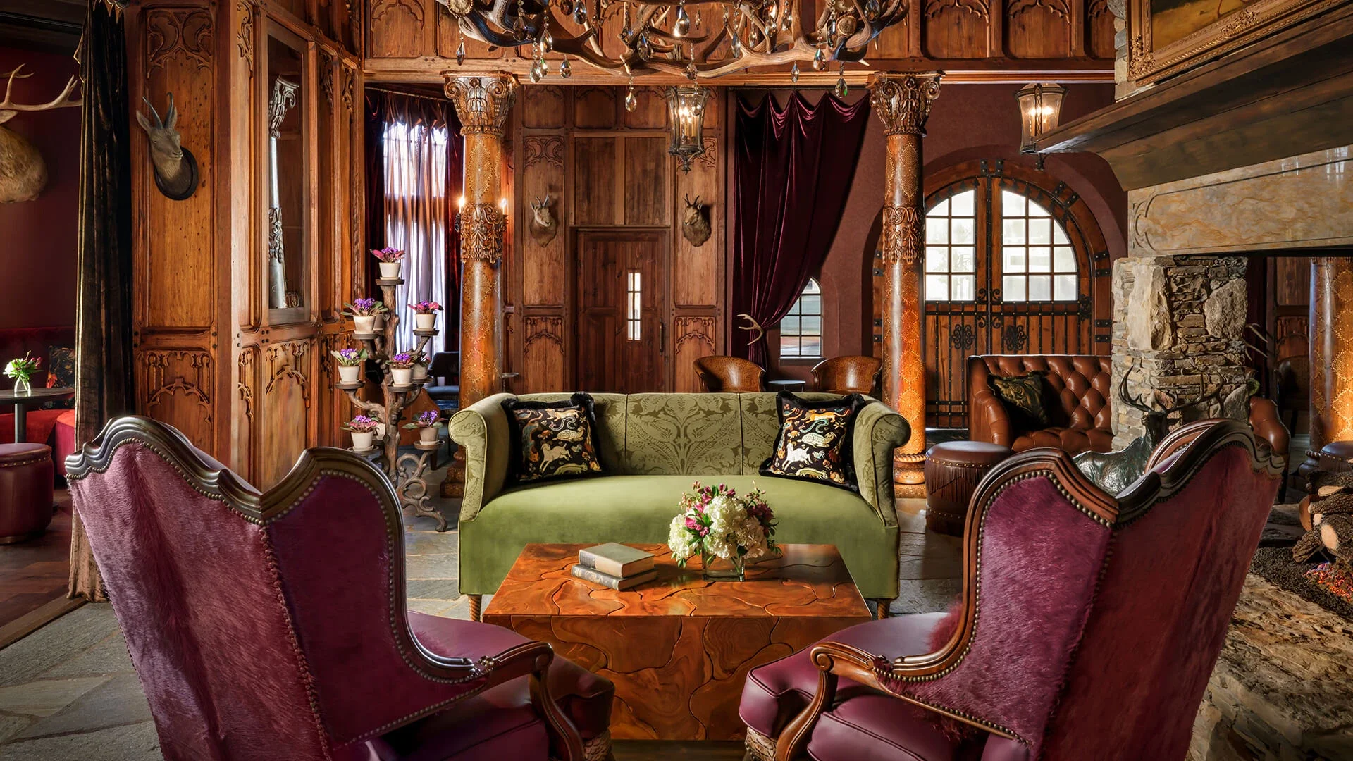 A luxurious, rustic living room with wood-paneled walls, ornate pillars, and plush furniture. There is a green sofa and pink velvet armchairs around a wooden coffee table with a bouquet of flowers. The room features deer head mounts, a stone fireplace, and large windows with red curtains.