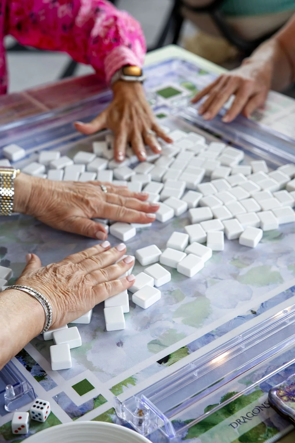 Multiple hands are seen playing a game of Mahjong with white tiles on a patterned mat.