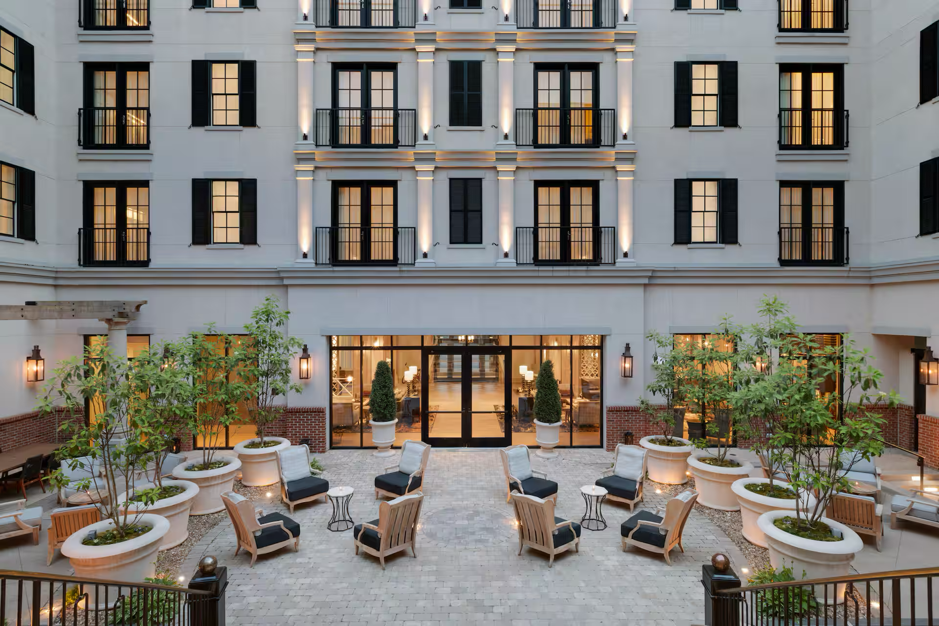 The image shows a multi-story building with lit windows and balconies, and an outdoor courtyard with seating and potted plants.