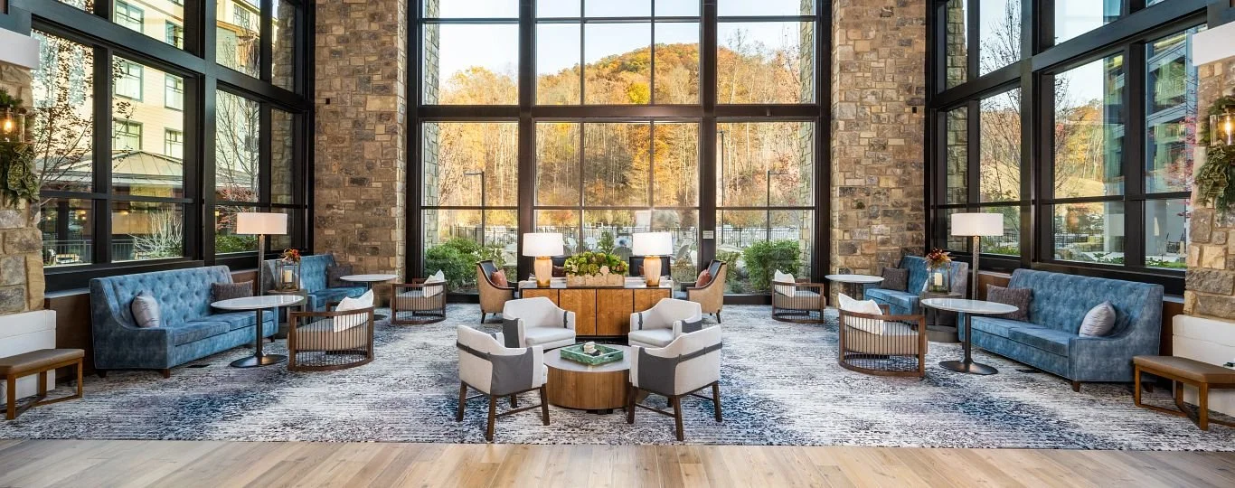 A spacious hotel lobby with large floor-to-ceiling windows showing autumn trees outside. The interior features blue velvet sofas, chairs, lamps, and a wooden sideboard with decorative plants, creating a cozy and modern atmosphere.