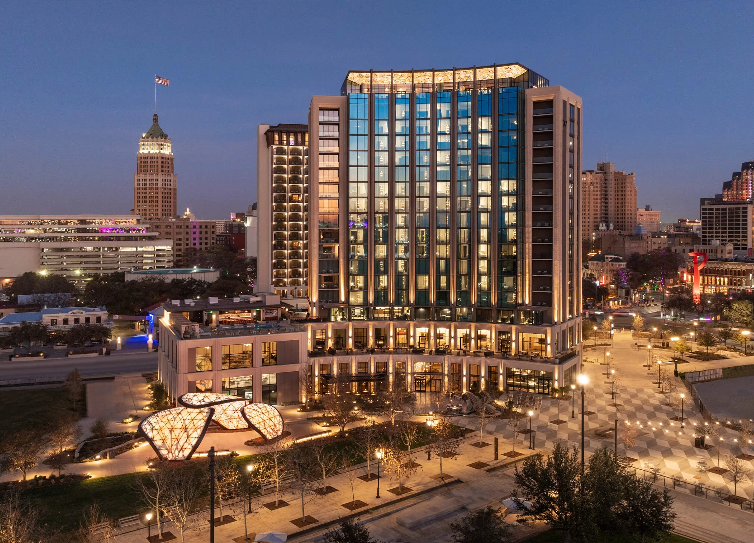 Nighttime cityscape featuring illuminated modern office building with glass facade, a historic clock tower building with an American flag, and a landscaped plaza with decorative lighting.