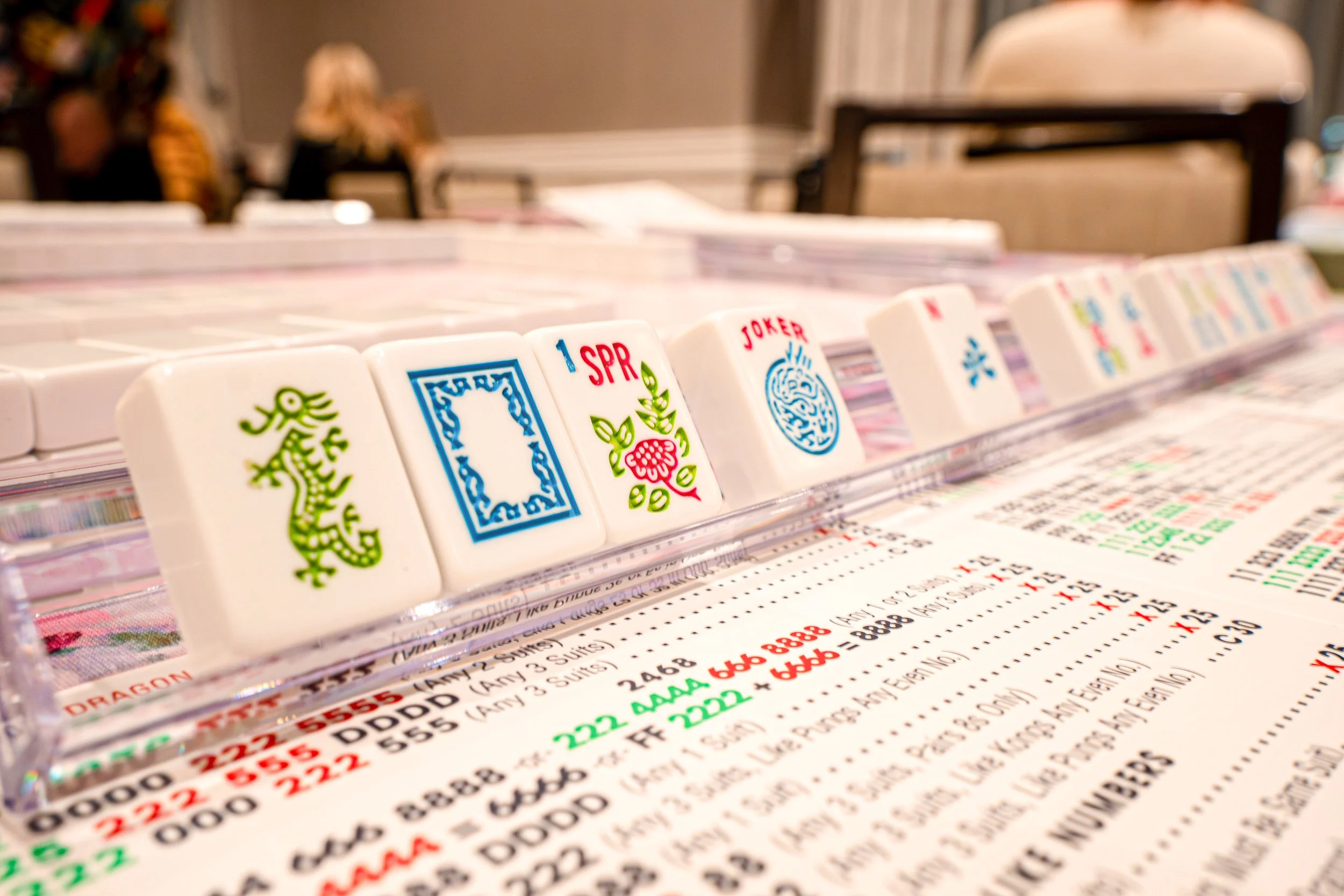Close-up of mahjong tiles with colorful designs, set up in a line on a printed sheet of paper with numbers and text, with a blurred background showing people sitting at tables.