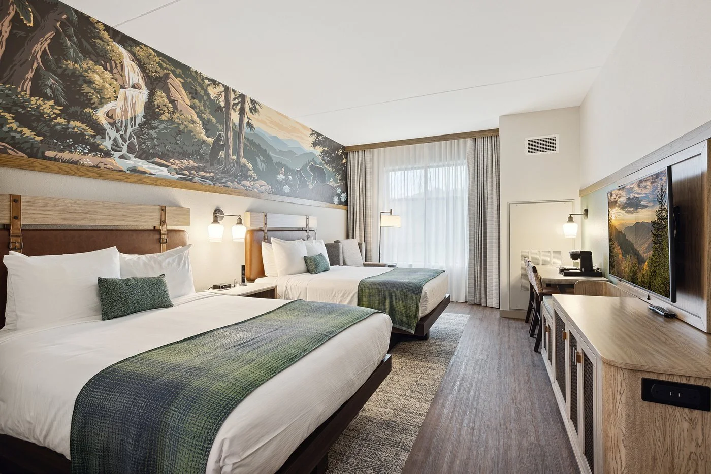 Hotel room with two double beds, a large nature mural on the wall, a desk with a coffee maker, and a flat-screen TV.