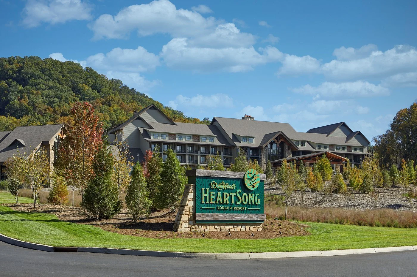 Lodge and resort named Dollywood's Heart Song Resort with a large building behind it, surrounded by trees and a hillside, under a partly cloudy sky.