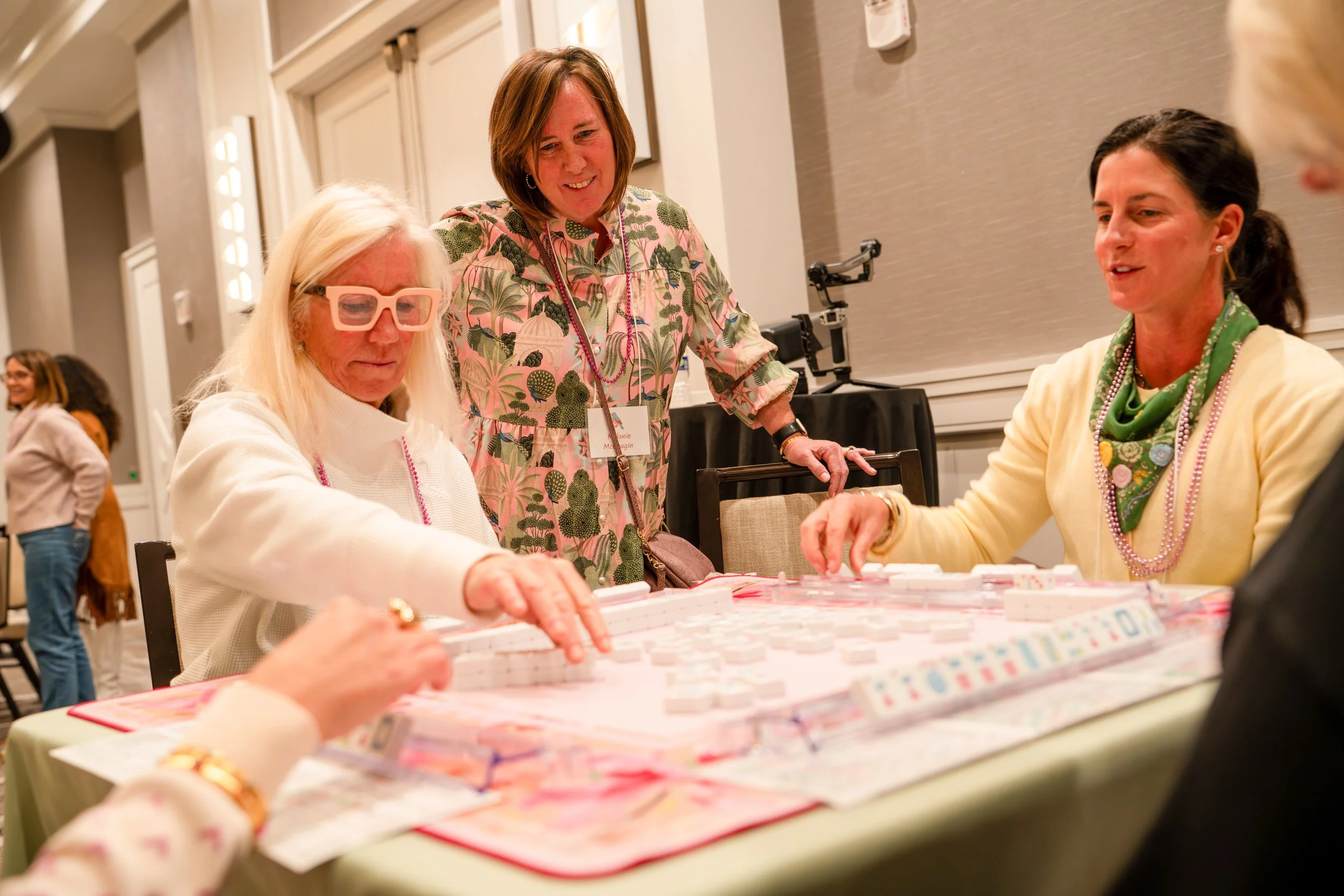 Four women are seated at a table playing a game of American Mahjo, with one woman standing and observing. The women are dressed in colorful clothing and have name tags. The setting appears to be a conference or social event in a large, well-lit room.