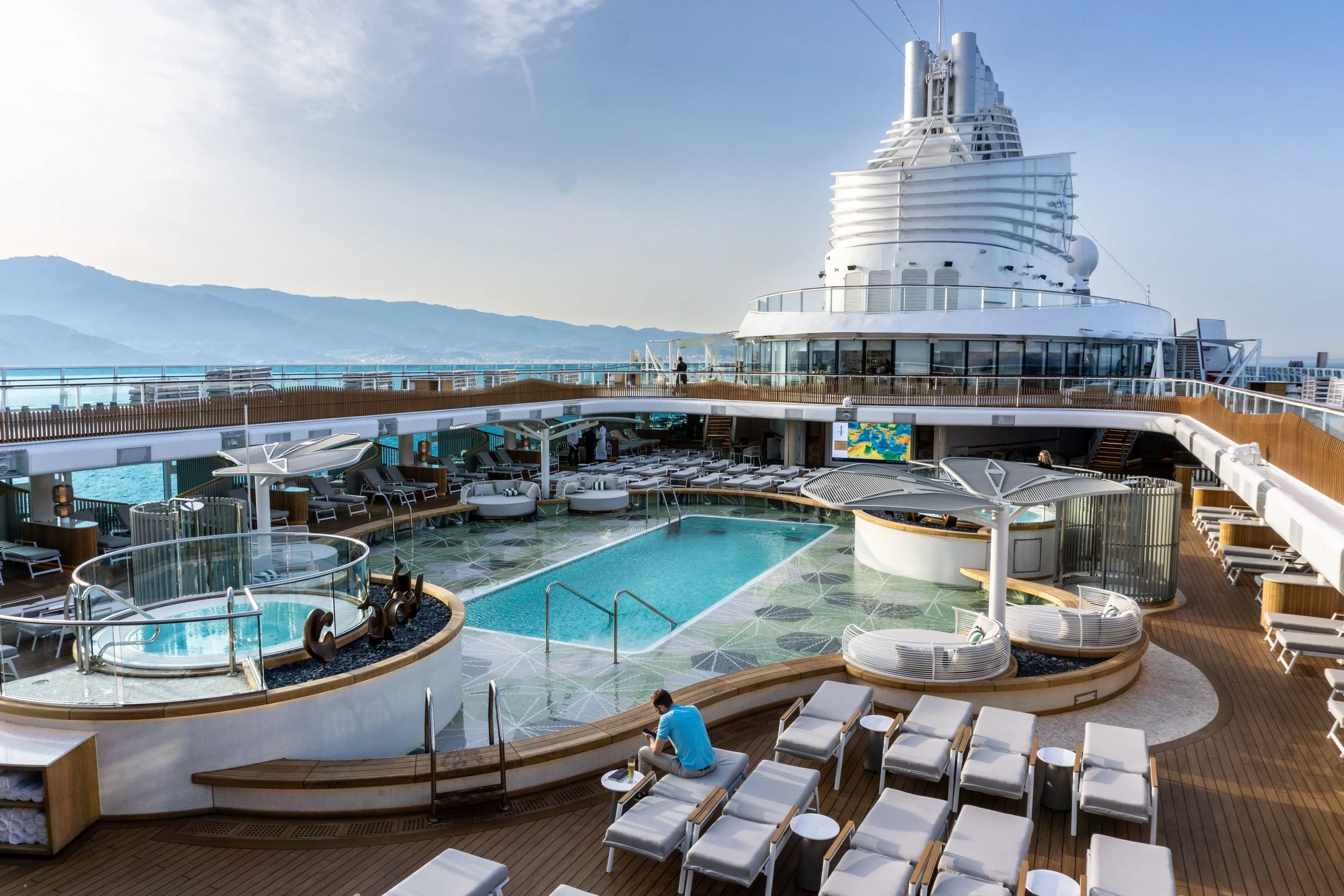 View of the top deck of a cruise ship with a swimming pool, lounge chairs, and a person sitting on a bench, with a mountain range in the background.