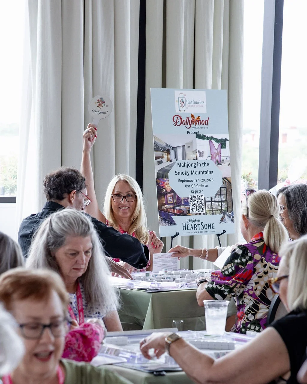 A woman at a mahjong event, holding up a fan with the word "Mahjong!" written on it. Several people are seated around tables, engaging in the activity, with a banner advertising a mahjong event at Dolly Parton’s HeartSong Lodge & Resort in the Smoky 