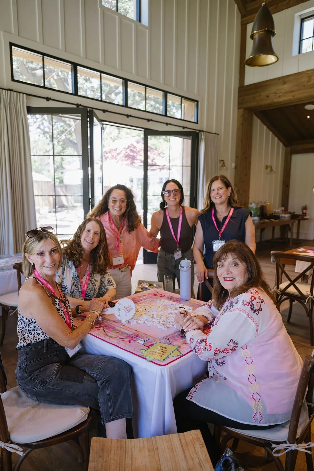 Group of six women gathered around a table at an indoor event, smiling for the camera. The table has name tags, a water bottle, and small items on it, with large windows and curtains in the background.