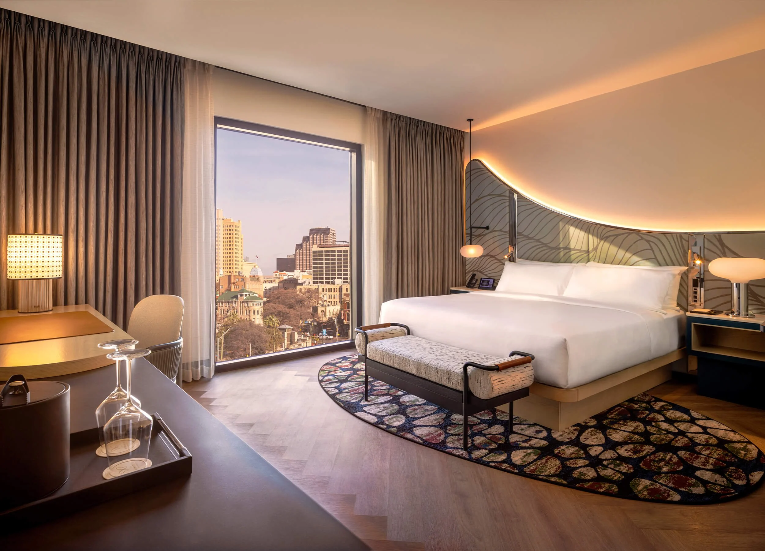 A hotel room with a large window showing city buildings outside, a king-sized bed with a decorative headboard, bedside lamps, and curtains.