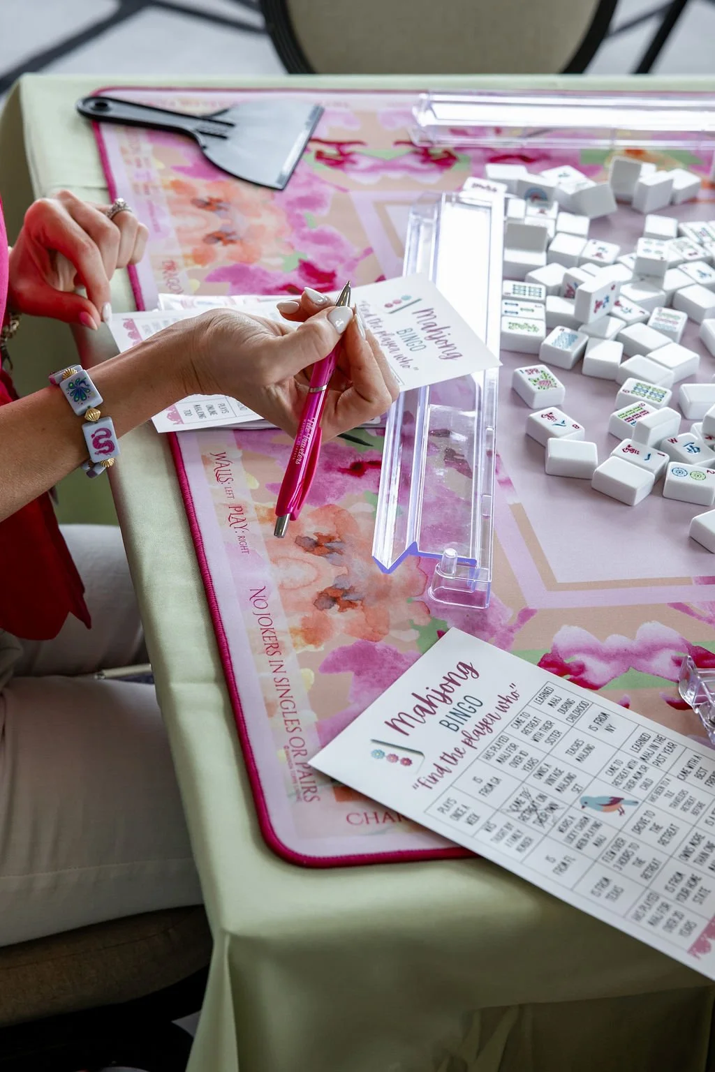 A person holding a pink pen and writing on a mahjong bingo card at a table with mahjong tiles decorated with a floral table cover.