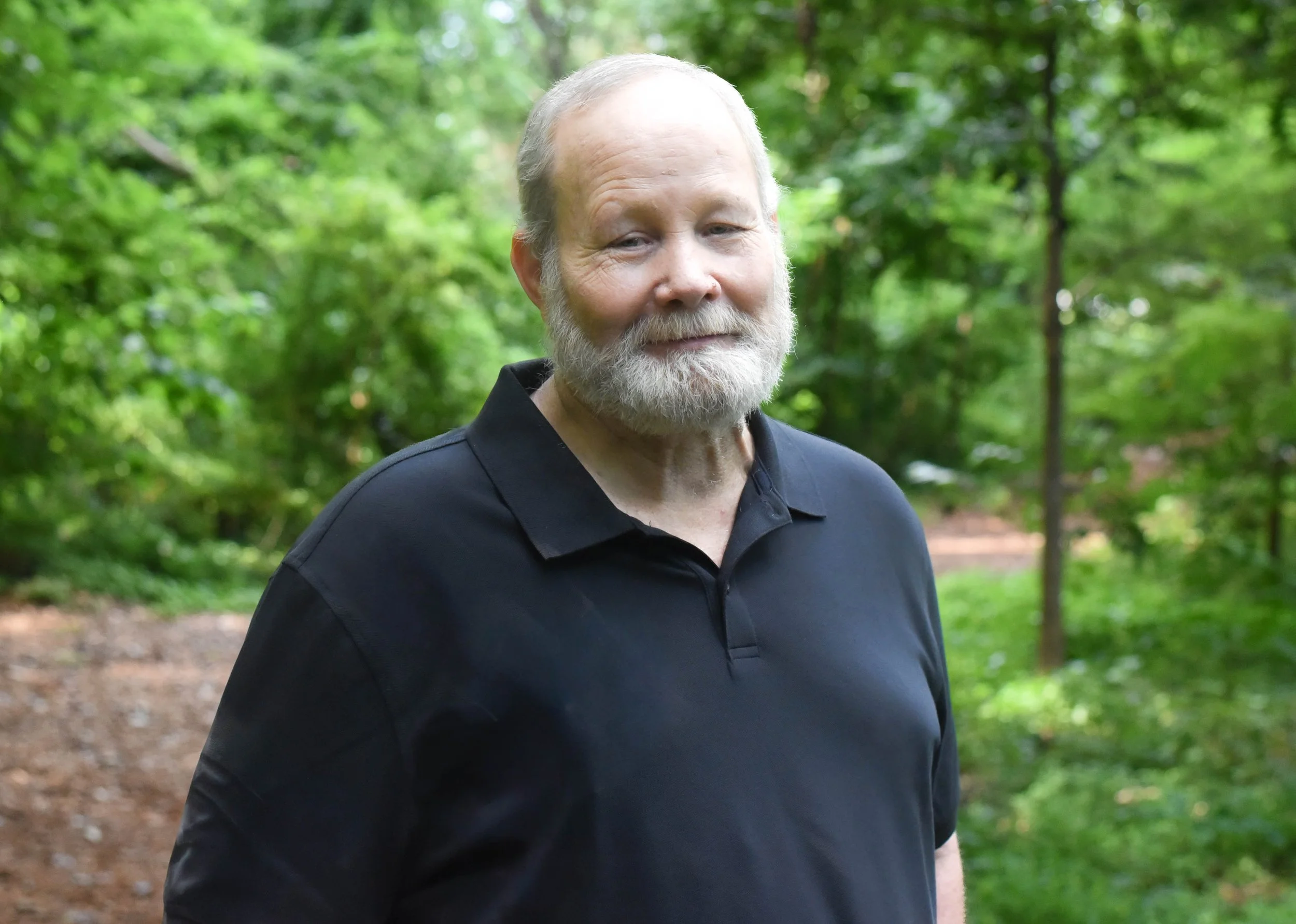 A smiling elderly man with a white beard and short hair, wearing a black shirt, standing outdoors in a green, wooded area.