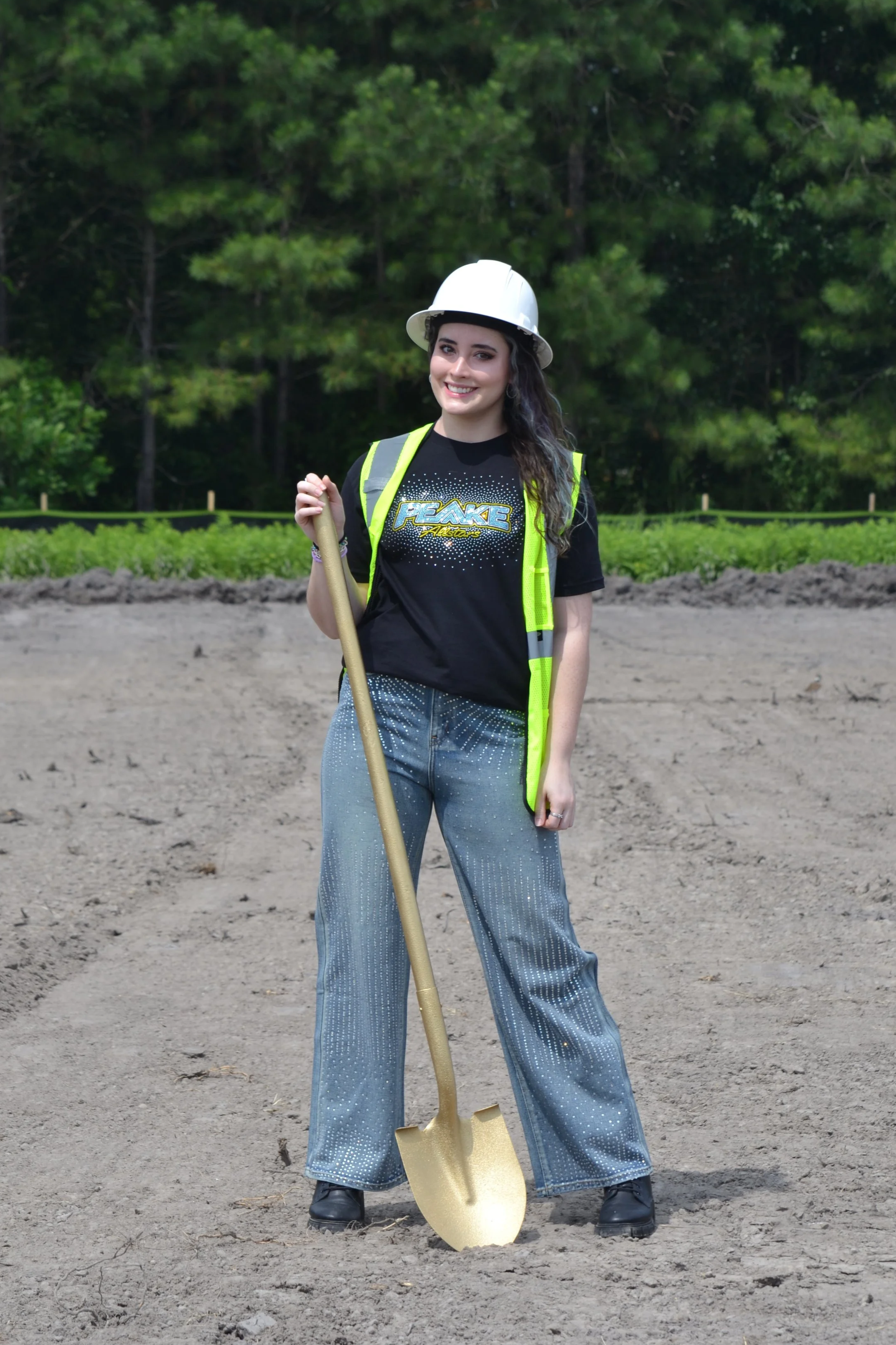 A young woman is standing outdoors on dirt, holding a shovel, wearing a white hard hat, a black graphic t-shirt, a neon yellow safety vest, blue jeans with a dotted pattern, and black boots. She is smiling and has dark, wavy hair.