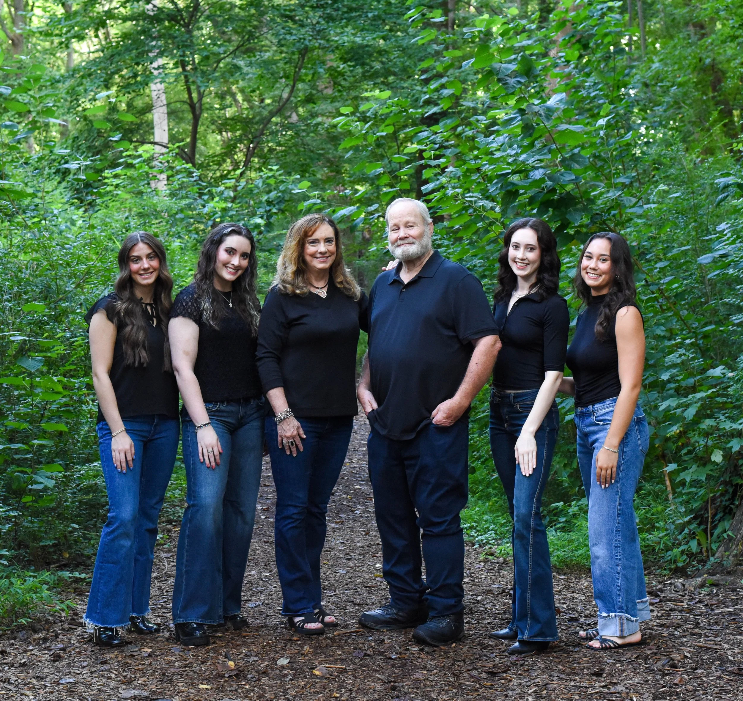 A family group portrait of six people standing on a forest trail surrounded by lush green trees and foliage. The group includes an older man and woman and four younger women, all smiling and dressed casually.