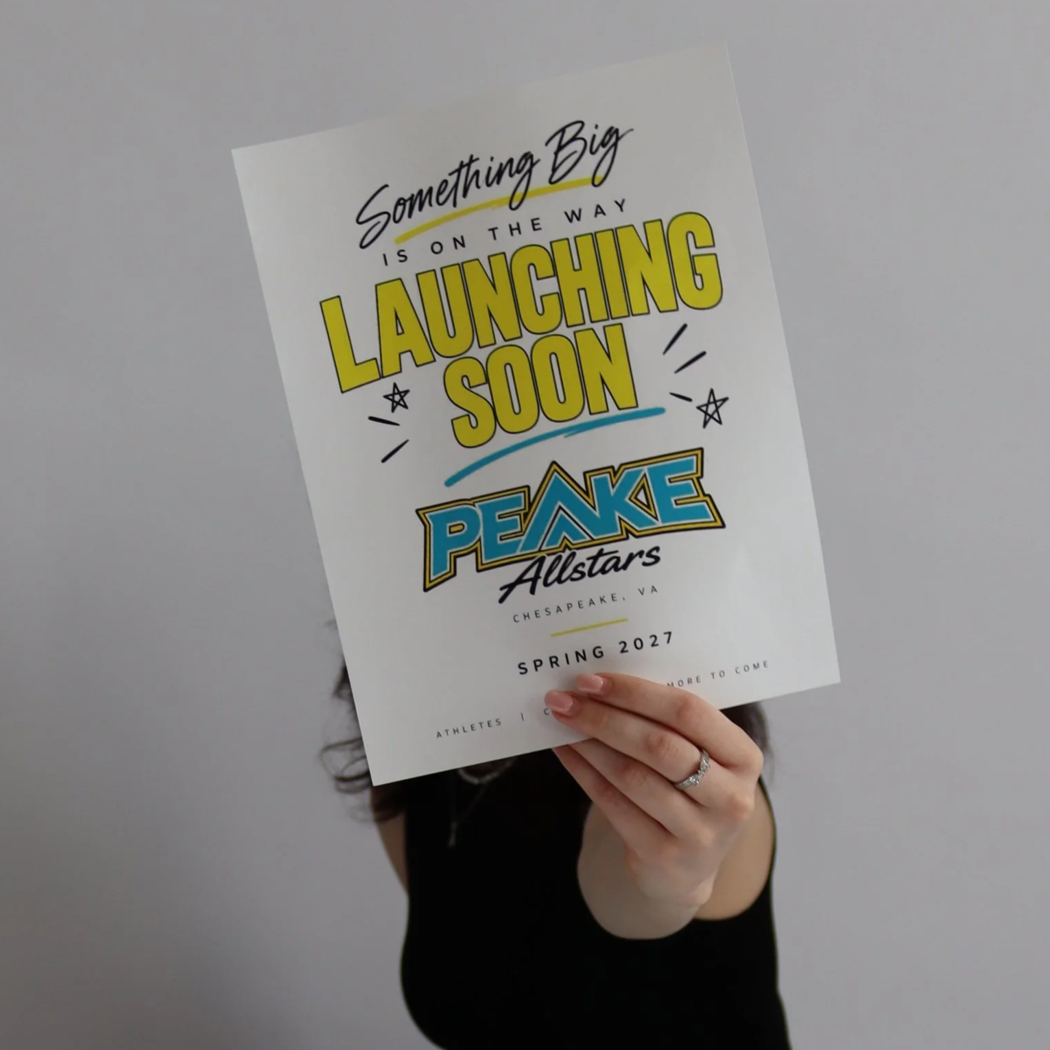 A person holding a poster that says 'Something Big is on the way Launching Soon PEAKE Allstars Chesapeake, VA Spring 2027' with a gray background.