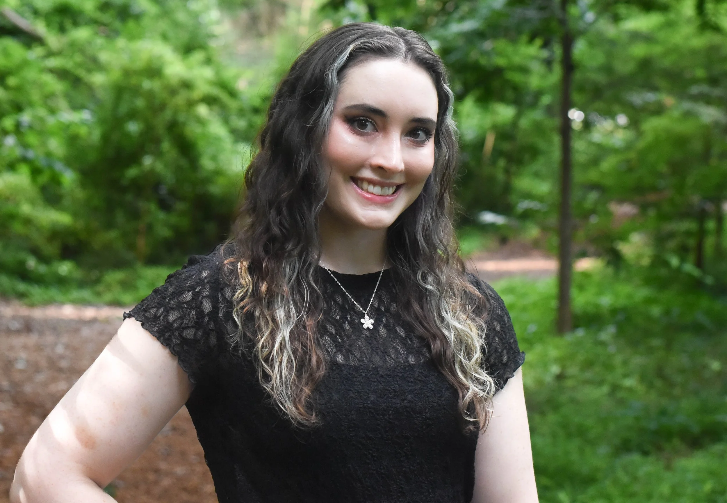 A young woman with long, wavy brown hair with gray highlights smiling outdoors in a wooded area wearing a black lace top and a flower pendant necklace.