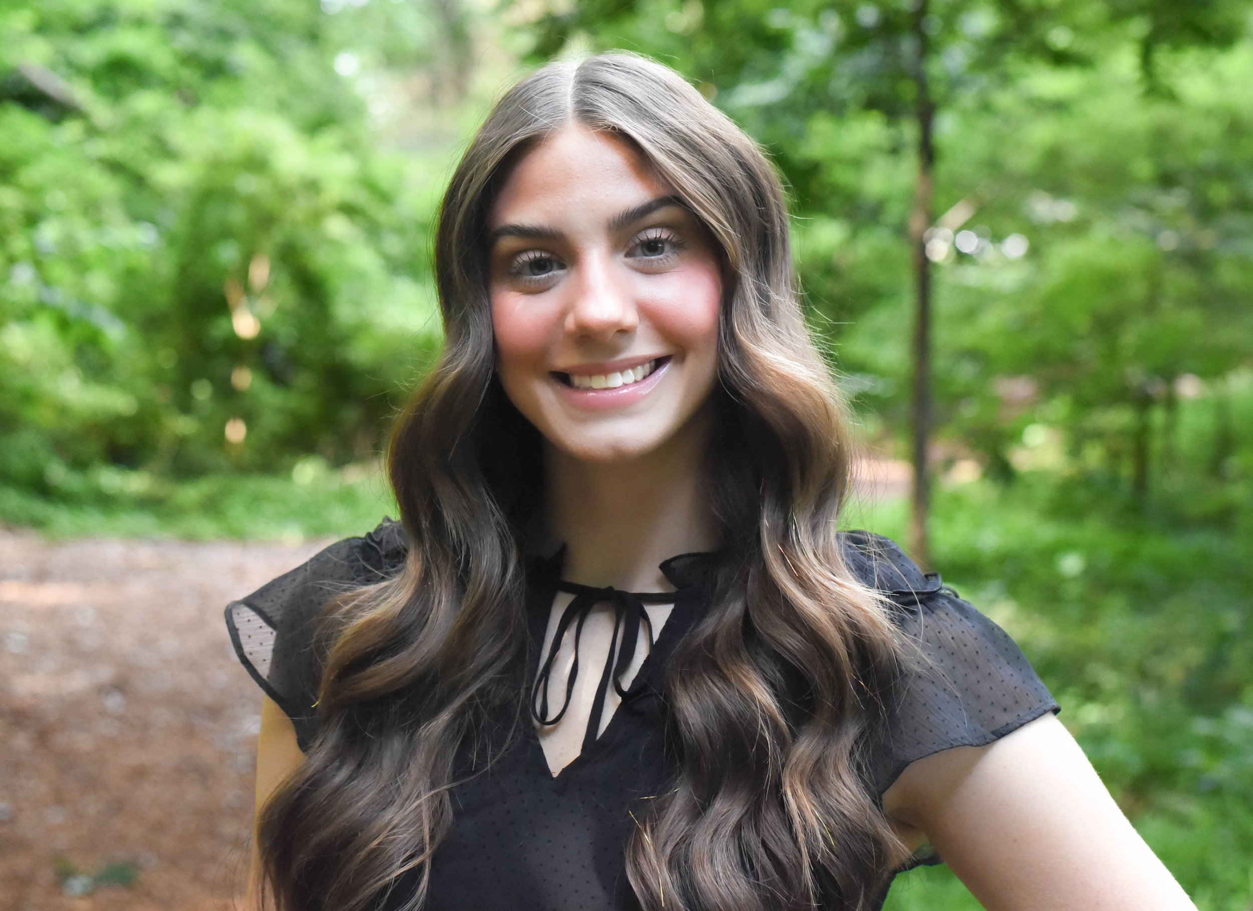 A young woman with long wavy brown hair and fair skin, smiling outdoors in a green, wooded area with blurred trees in the background.
