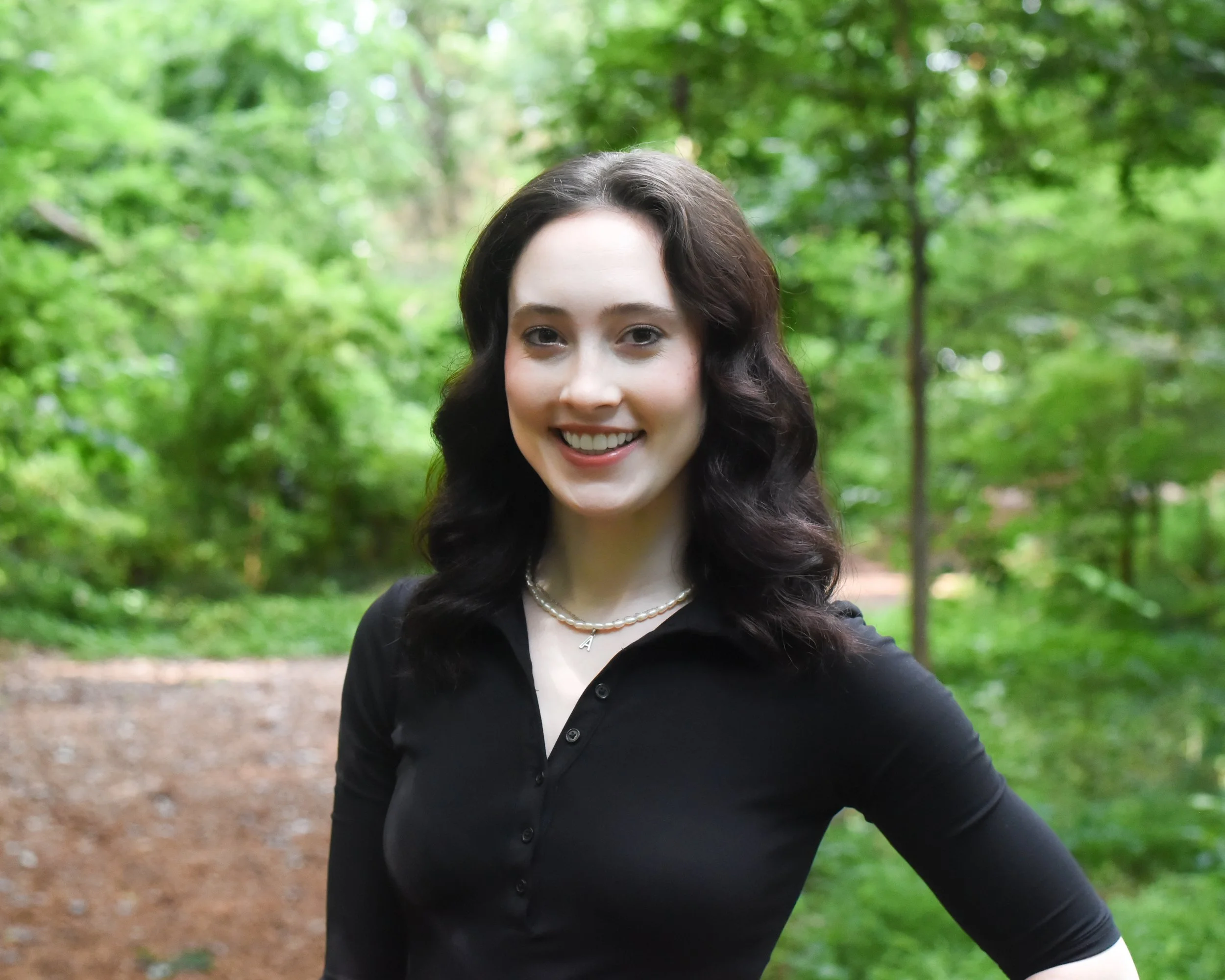 Portrait of a young woman with dark hair smiling outdoors in a green forest during daytime.