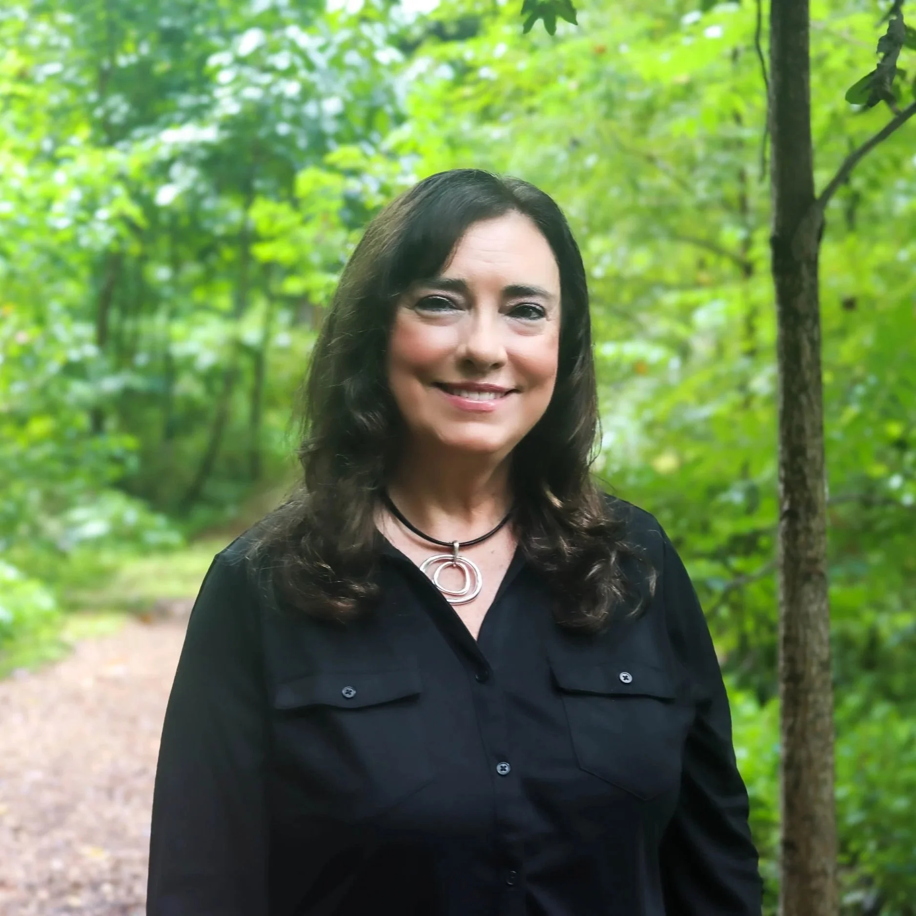 A woman with dark hair smiling outdoors in a green forest setting with trees and a dirt path.