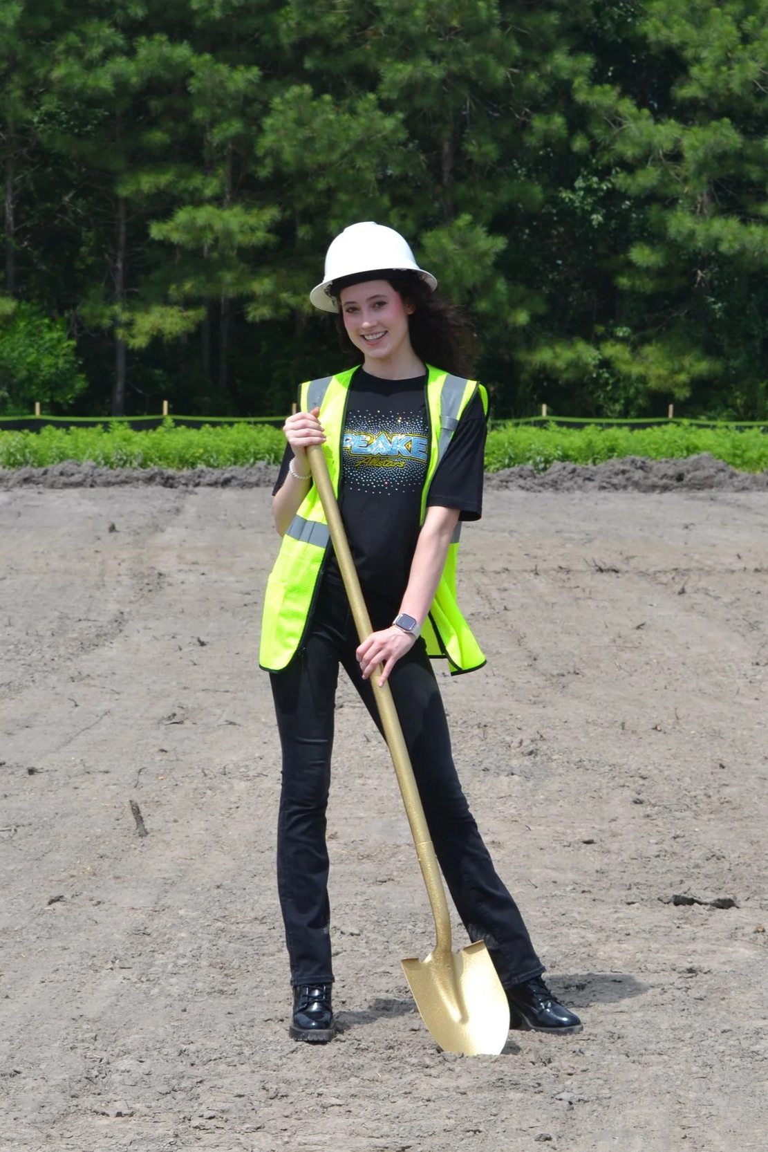 A young woman wearing a white hard hat, a yellow safety vest, and black clothing, holding a golden shovel at a construction site with a background of green trees and soil.