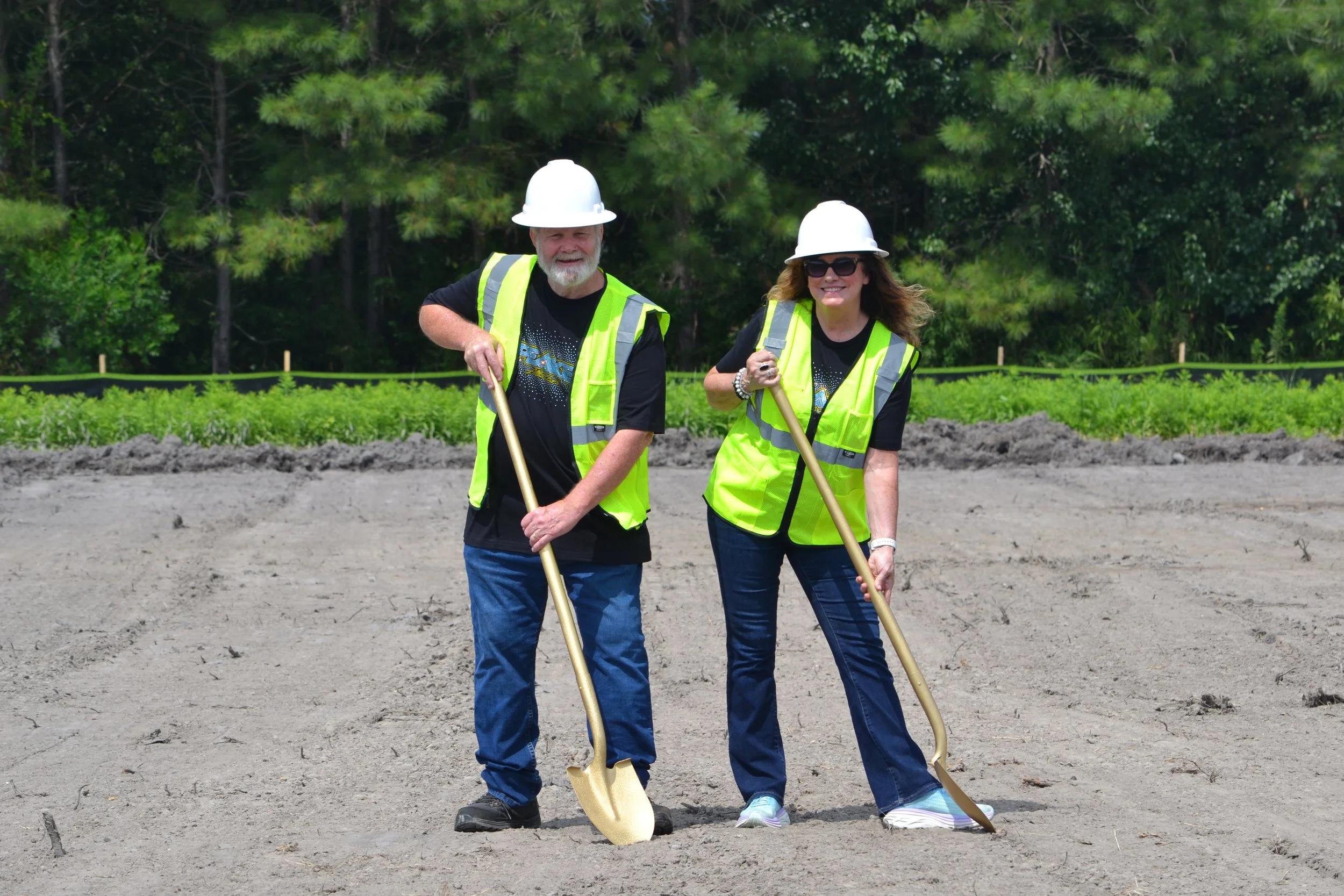 Two people, a man and a woman, wearing hard hats and reflective safety vests, are standing outdoors on a construction or landscaping site with trees and greenery in the background, holding shovels.