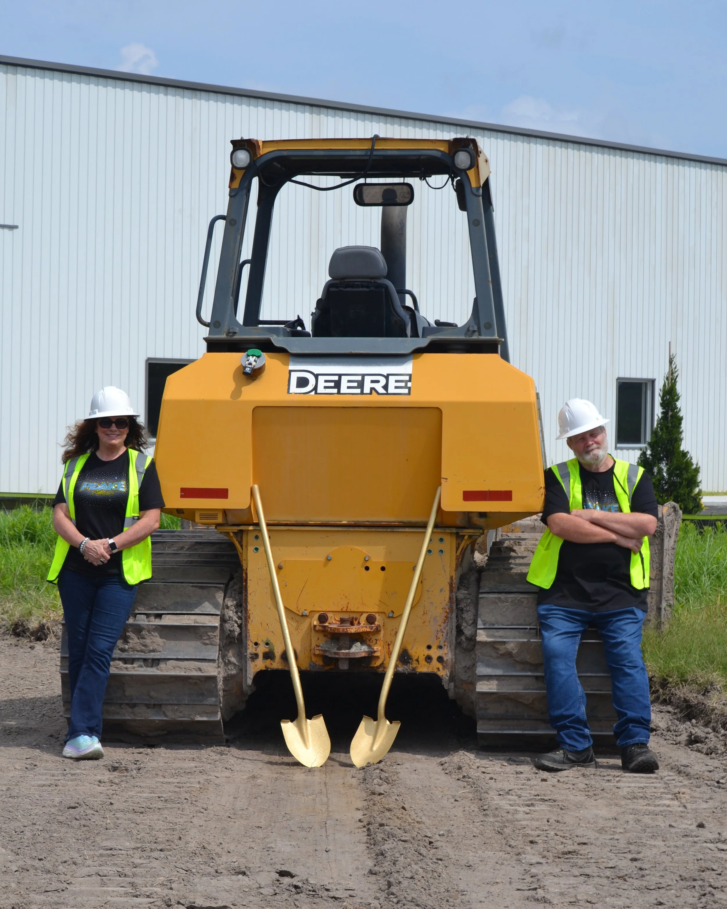 Two people standing next to a large yellow John Deere bulldozer outdoors on a dirt construction site, wearing hard hats and safety vests, smiling and posing for the photo.