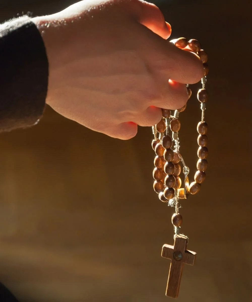 Close-up of a hand holding a wooden rosary with a wooden cross, backlit to highlight the beads and cross.