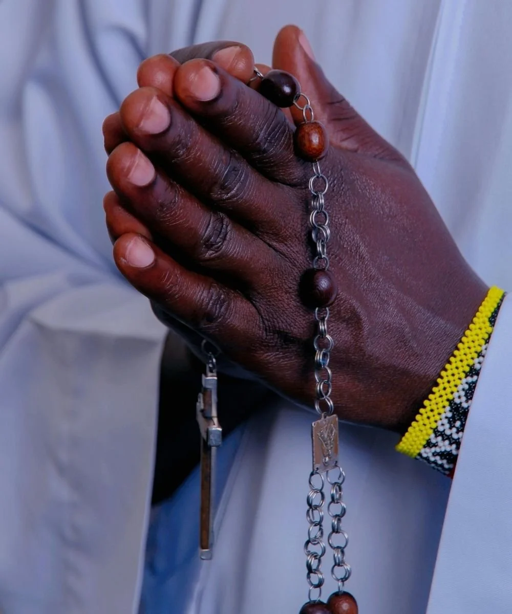 Person holding a rosary with dark and light wooden beads and a metal cross, wearing a colorful beaded bracelet.