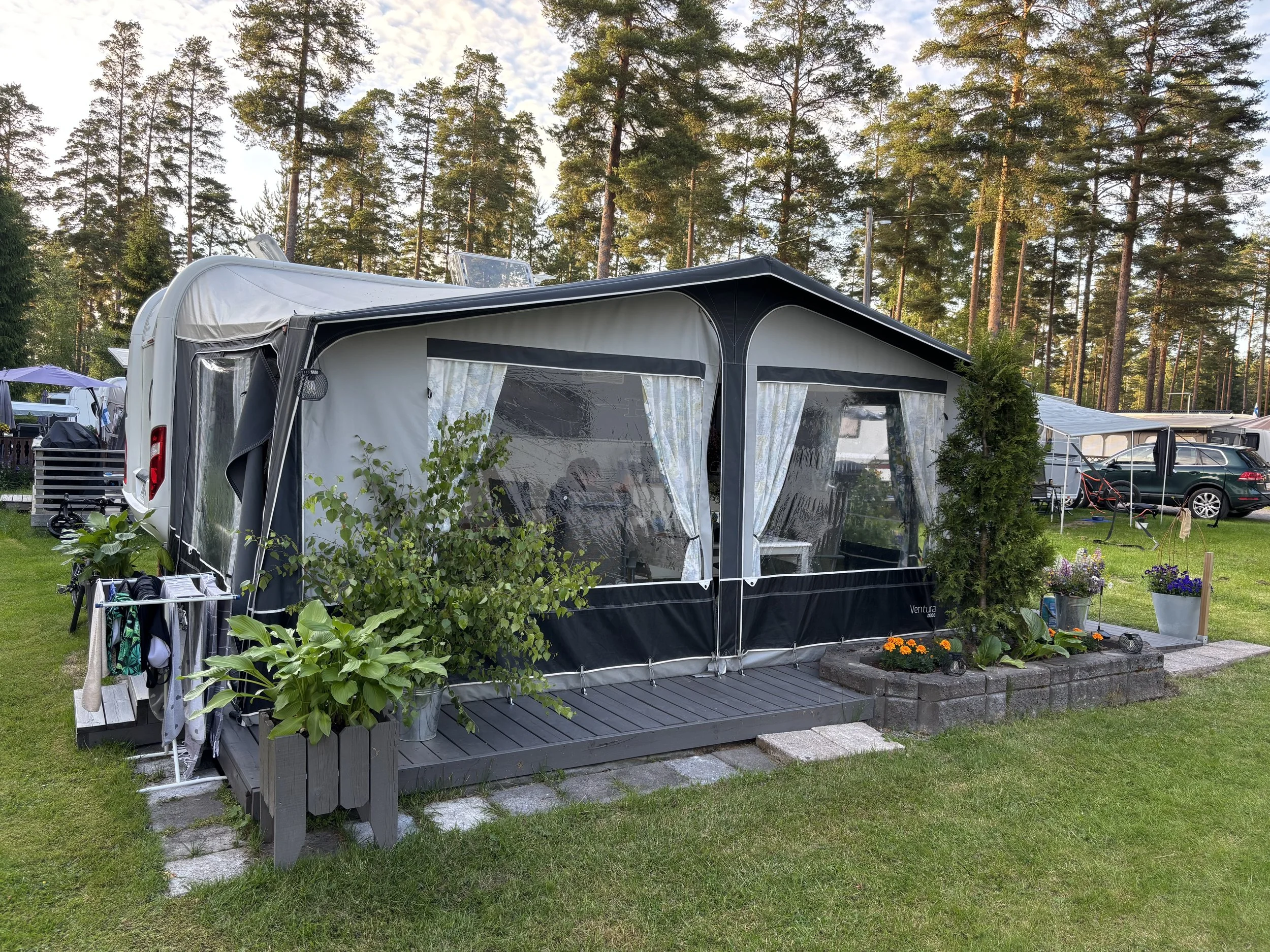 Vaalea matkailuauto tai asuntoauto, osa ulkosalla, taustalla puita ja muuta matkailualueen rakennuksia.