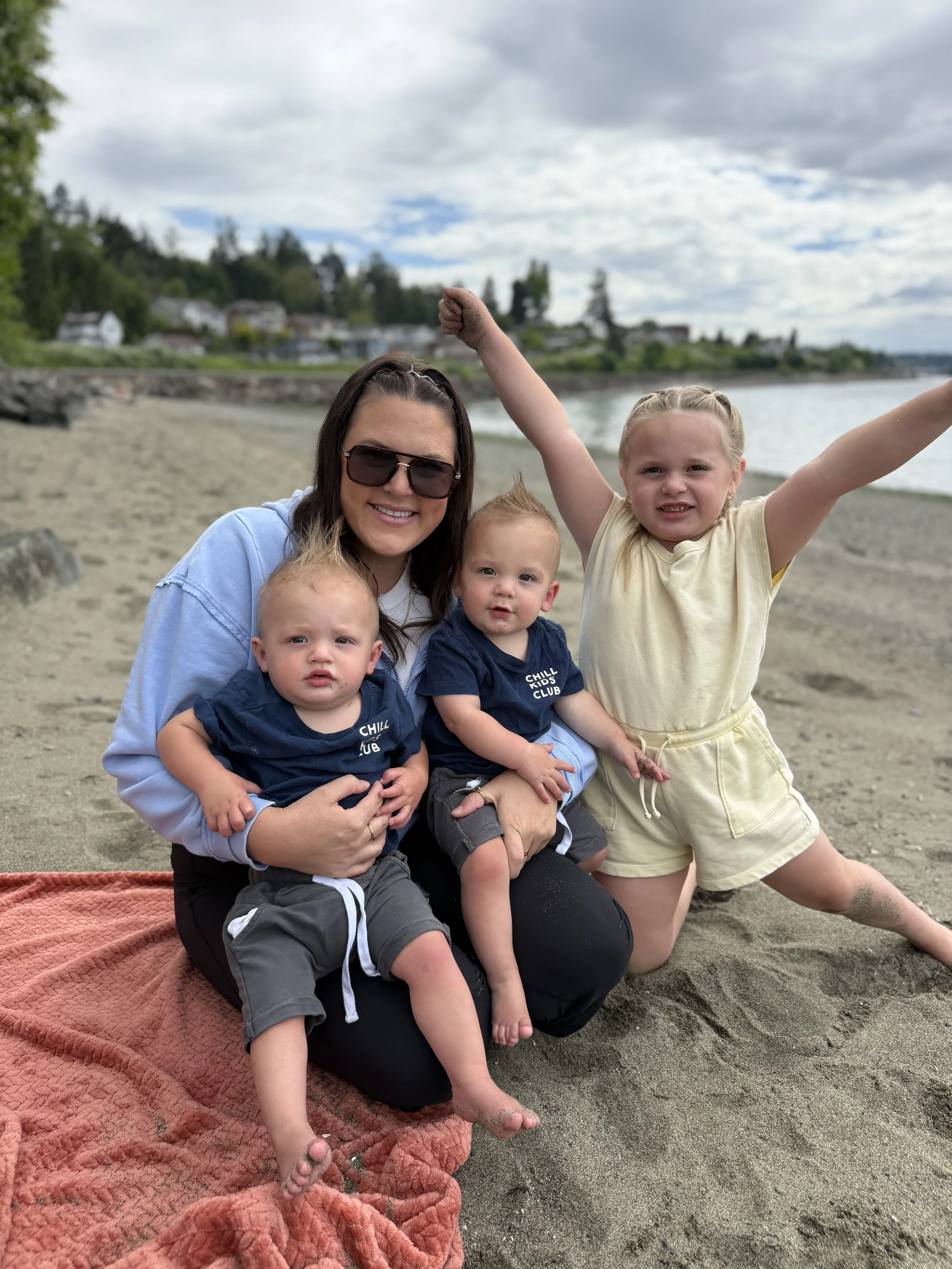 A woman and three young children sitting on a beach with a body of water and houses on a hill in the background. The woman is wearing sunglasses, a light blue sweatshirt, and smiling. The children are dressed casually, with two wearing matching navy blue shirts that say 'CHILL KIDS CLUB' and one girl in a light yellow dress, raising her arms in the air.